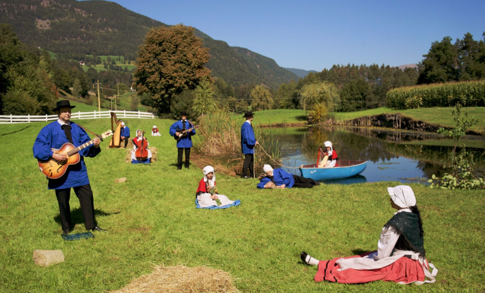 10 figures in folk clothing and musical instruments, standing in a grassy field by a small lake