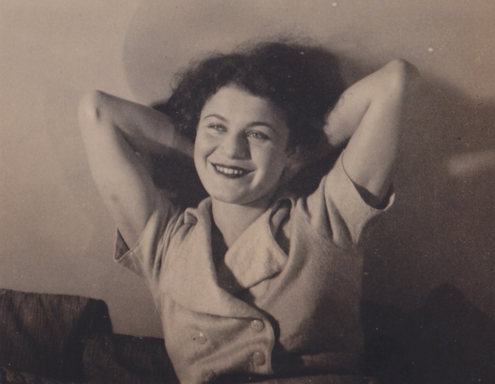 Photograph of a woman (Renee Gross) who faces the viewer. Her bent arms are raised, and her head leans on them, against a wall behind her. She is looking and smiling at someone who appears to be to the left of the viewer.
