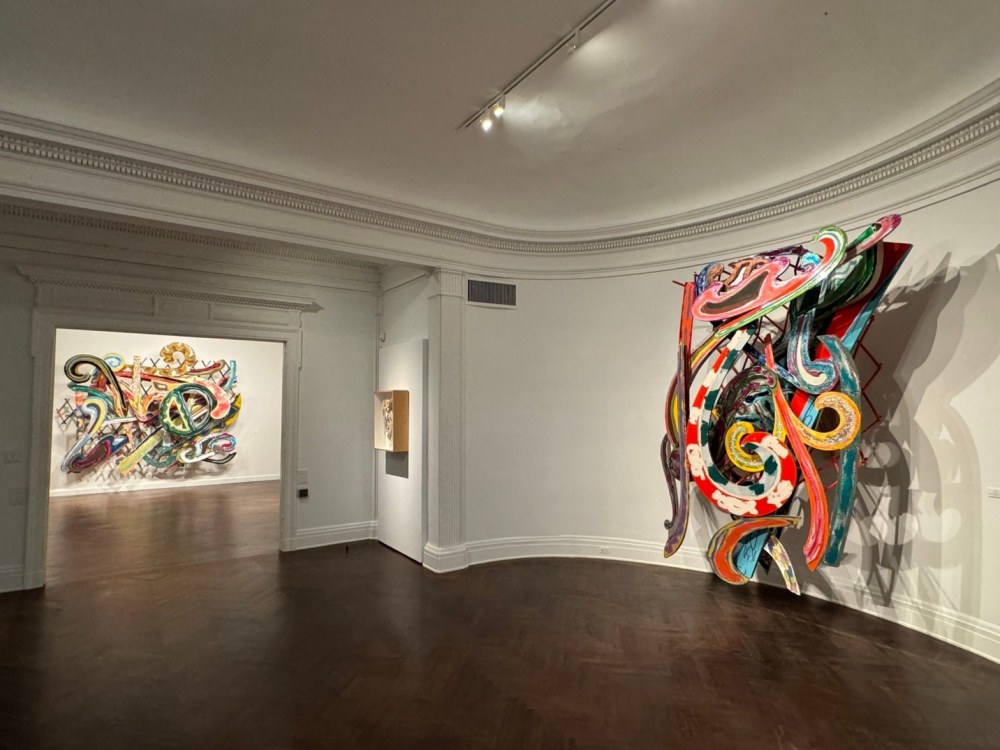 Bright colored, swirling sculpture-paintings hanging in a white room with dark wood floor