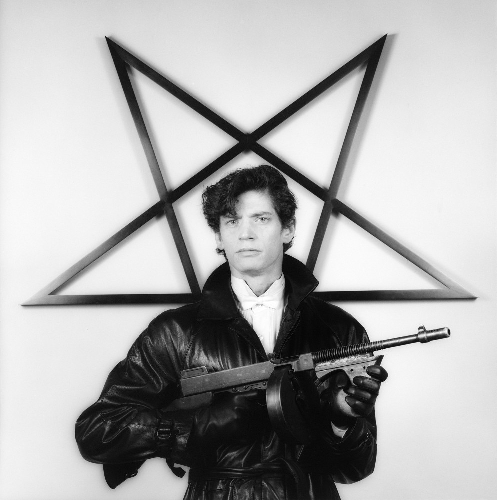 Robert Mapplethorpe wearing a leather jacket and gloves and holding a gun in front of a star sculpture.