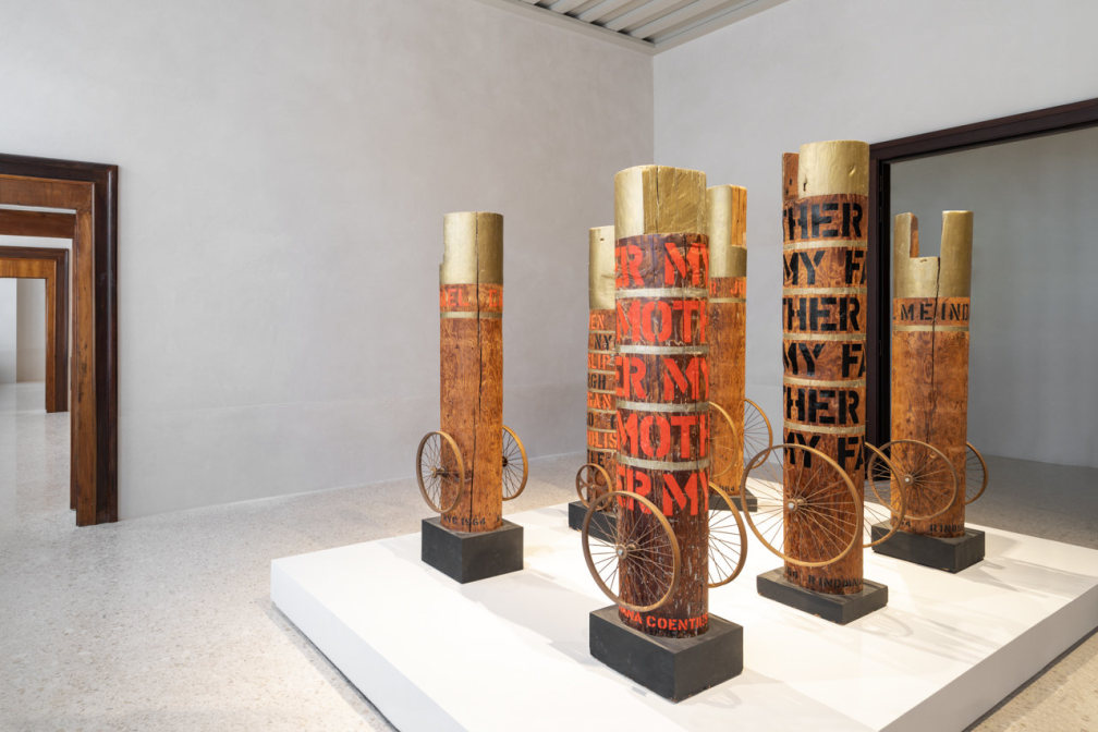 Installation view of six columns, each with red or black writing, gold tops, and wheels.