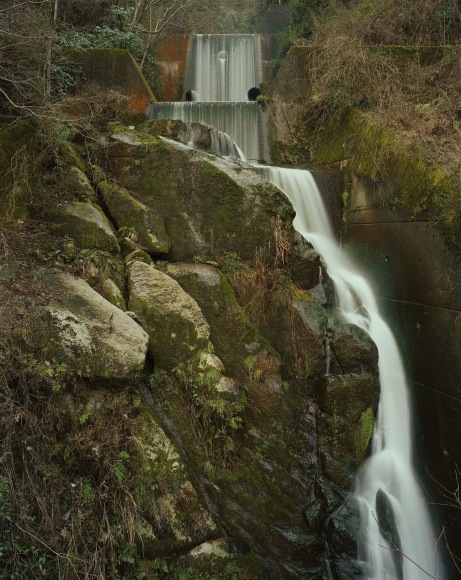 Color photograph of water spillage cascading from a dam in a wooded area.