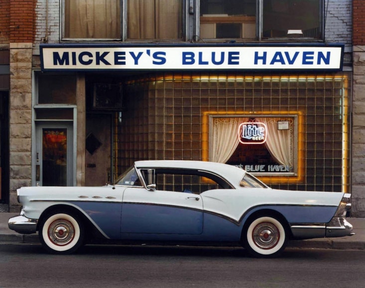 Color image a blue classic 1950s car in front of a working g class bar with a sign that reads Mickey's Blue Haven.
