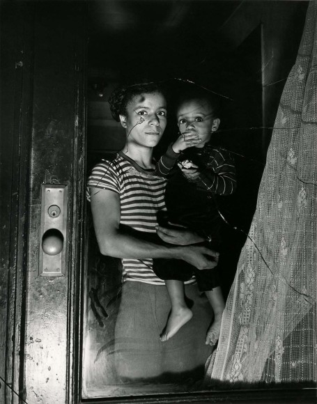 Black and white image of a mother and child, looking at the camera through a broken window in Harlem.