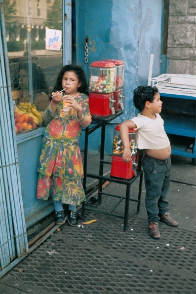 Color photo of two children hanging around outside an urban shop by a gumball machine.