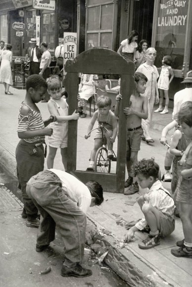 Black and white photo of a mid-20th century street scene showing a group of young boys surrounding the frame of a brown mirror on a curb. A child on the tricycle looks through the frame.