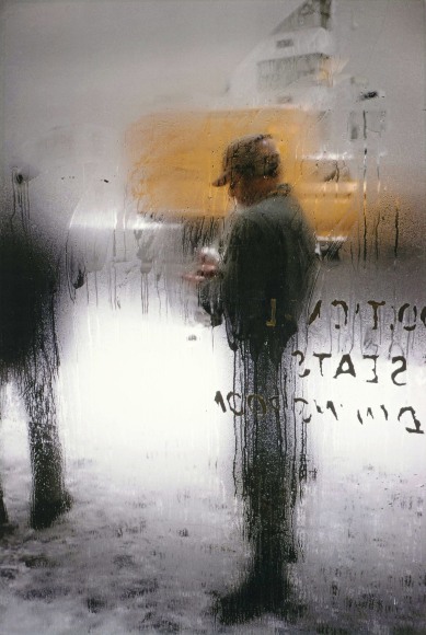 Color photograph of the blurred image of a man on a snowy street, scene through a window dripping with condensation.