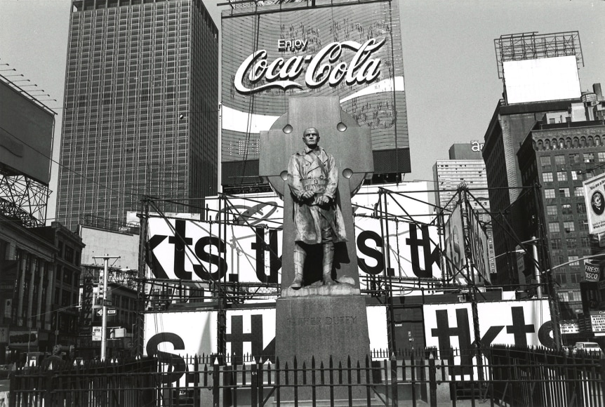 Black and white photo of a bronze statue of military Chaplin Father Duffy, standing in front of a cross, with a hidpodge of urban signing behind him. One prominent sign over his heads reads&quot; &quot; Enjoy Coca-Cola&quot;
