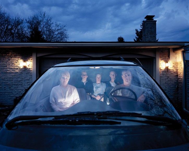 Blue toned photographic portrait of a suburban family in a mini van, taken through the windhshield.