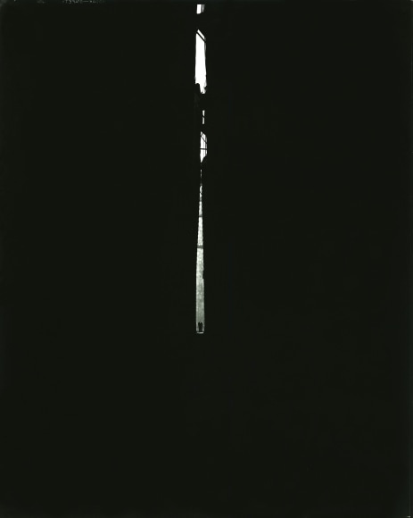 Nearly all black photo showing a shaft of light in the center, framing a man seen in the distance.