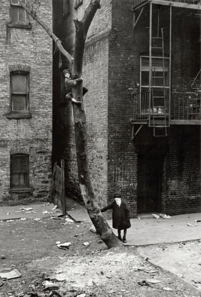 Black and white photo showing tow kids in homemade masks posing for the camera, one is leaning on a tree that the other has climbed up.
