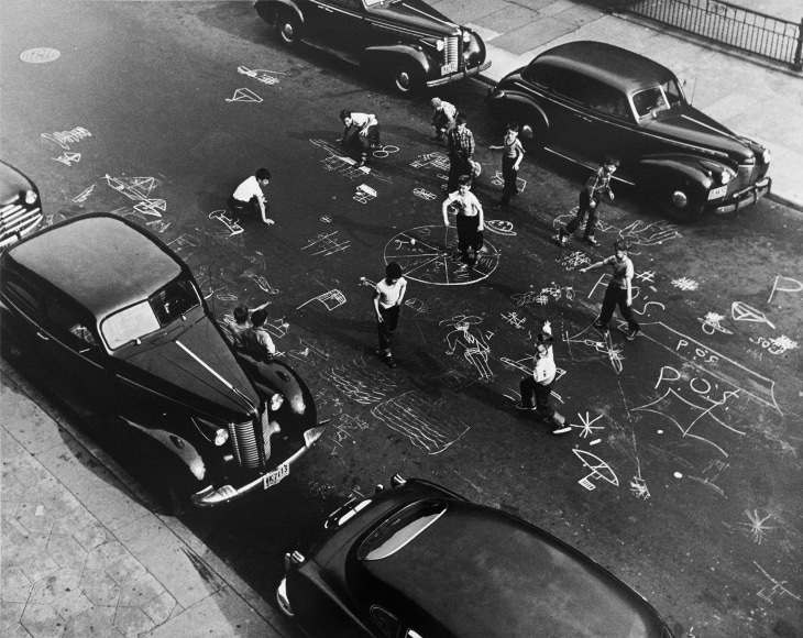 Black and white photo from above of children drawing all over a NYC street in the 1950s, with mid-century cars parked ont he street.