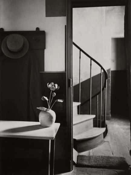 Black and white photo of a flower vase on a table in front of a coat and hack on a rack, with a spiral staircase behind.