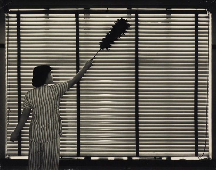 Black and white photo of a cleaning person dusting a set of gentian blinds that resembles her striped uniform.