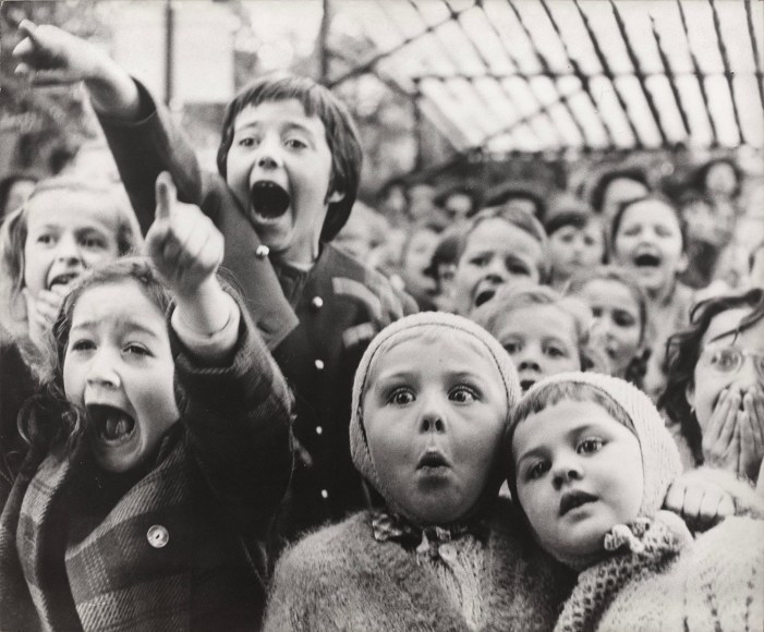 Black and white photograph of children's expressions of surprise and excitement as they watch from the audience of an outside performance.