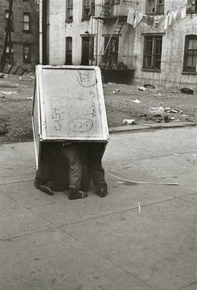 Black and white photo showing a large wooden box in an urban neighborhood, with legs poking out, revealing that children are playing inside.