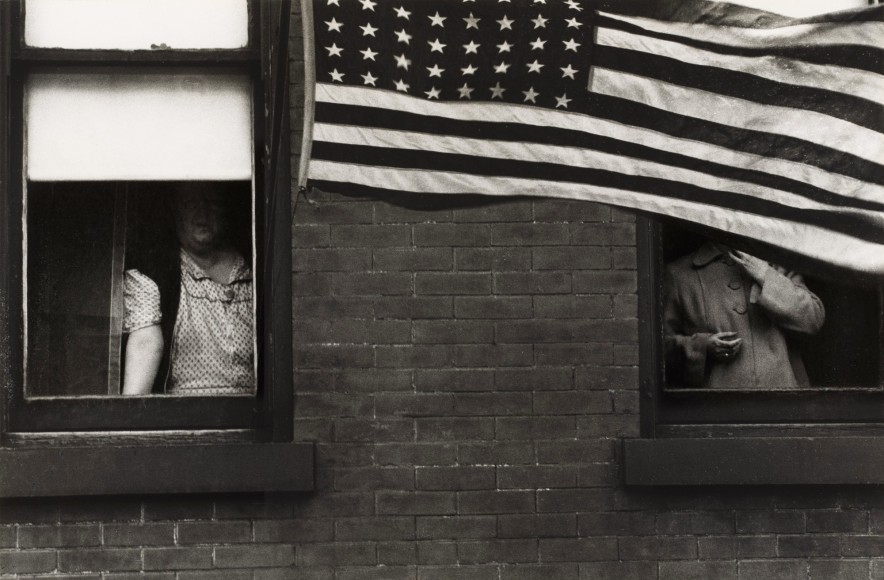 Black and white photo of a person looking out of a brick apartment building's window, their faces totally obscured by the shadow of a window shade and a waving American flag.
