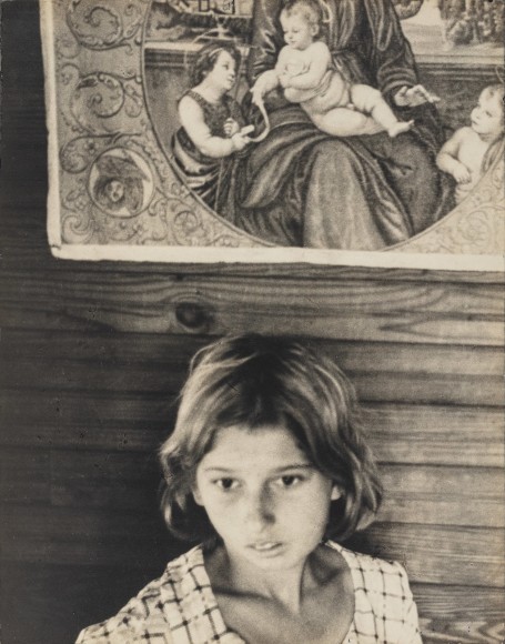 Early 20th entry photographic portrait of a young girl in. applaud dress with downcast eyes, in front of a clapboard was with a print of the Madonna and Child on it.