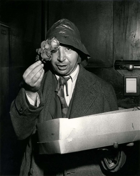 Black and white image of a flower seller holding flower up to the camera. Wearing a slouchy hat with a box.