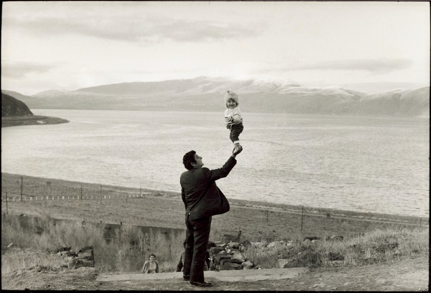 black and white photo of a man in a suit holding a small child up with one hand in front of a large mountain lake.