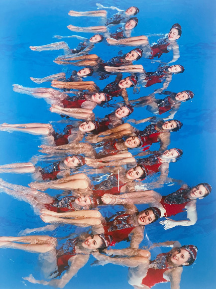 Color photo of a team of synchronized swimmers posing for the camera in a swimming pool.