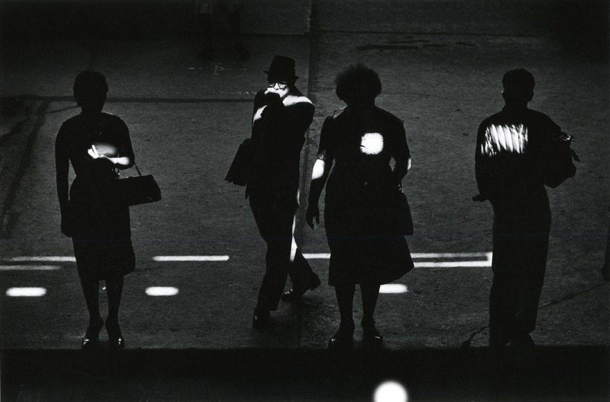 Black and white photo of pedestrians standing in shadow with spots of light falling on them.