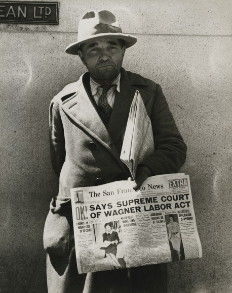 Black and white photo of a newspaper dealer with a copy of the San Francisco News from 1937 announcing Supreme Court decision on Wagner Labor Act