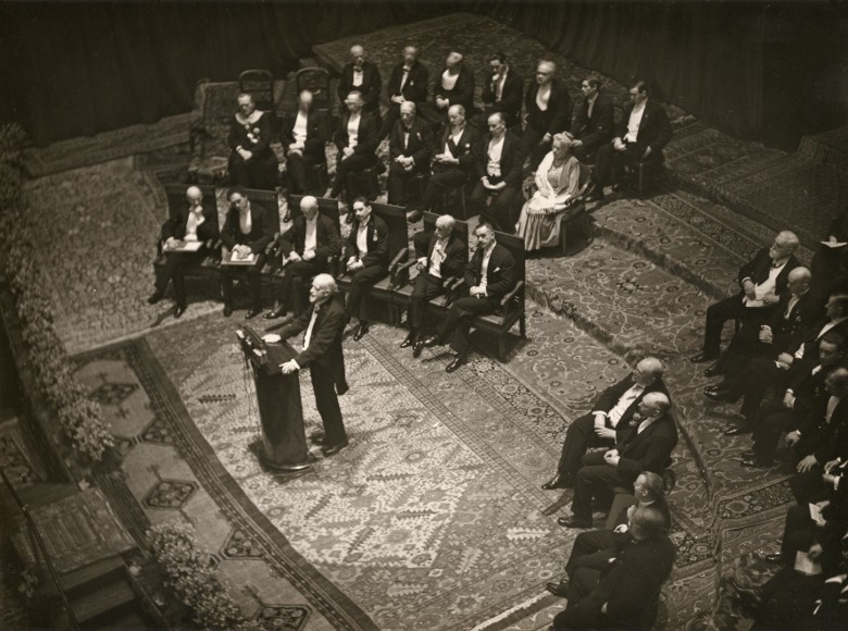 Black and white photo of the 1929 Nobel Prize ceremony&mdash;a grey bearded man speaks at a podium, while a formally dressed audience sits behind him.