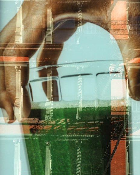 Color photograph of a hand holding a glass of green juice superimposed as a reflection on the facade of a NYC building.