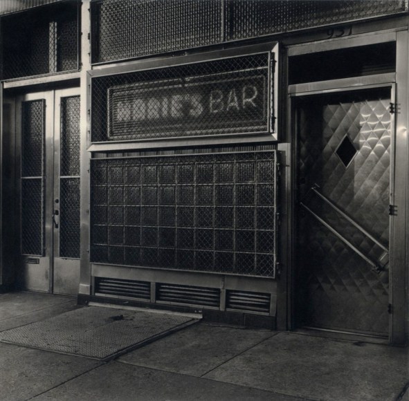 Black and white of a neighborhood bar with windows covered in protective e metal grating, a neon sign says &quot;Marie's Bar&quot;.
