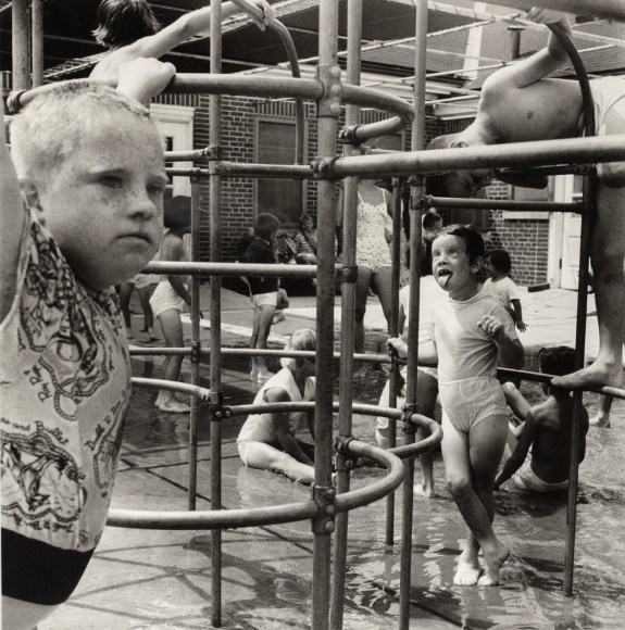Black and whit photo of kids playing on monkey bars. One girl sticks her tongue out.