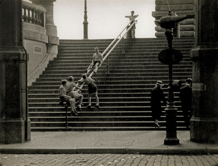 Black and white photo of a group of children watching their friend slide down the handrail on large public stone steps.