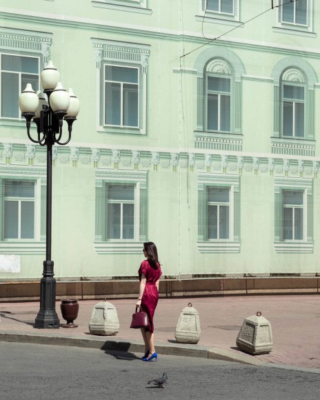 Woman in a red dress and blue heels standing with her back to the camera in front of a life-size printed facade of a building.