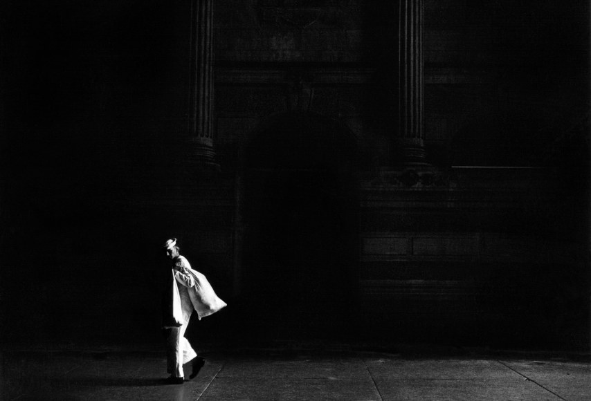 Black and white image of a sailor carrying a knapsack, illuminated by strong slanting light abasing a dark background&mdash;with a classical revival column is faintly seen in the darkness behind him.