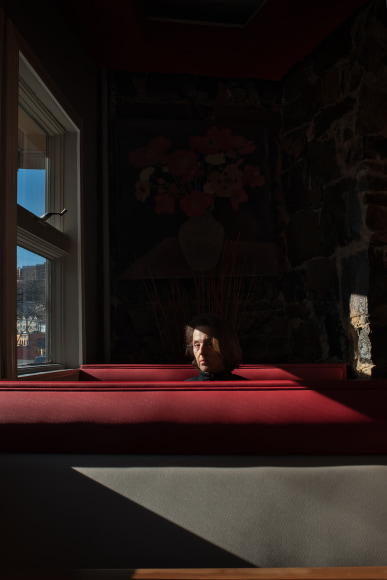 Woman sitting alone in a diner booth, next to a window, in slanting light. She is looking at the camera.