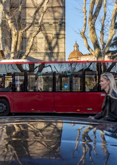 View of a street in Rome, lookin across the street at an old Jewish Temple, with the top of a car in the foreground, reflecting trees, with a female cyclist and a red bus passing on the street.