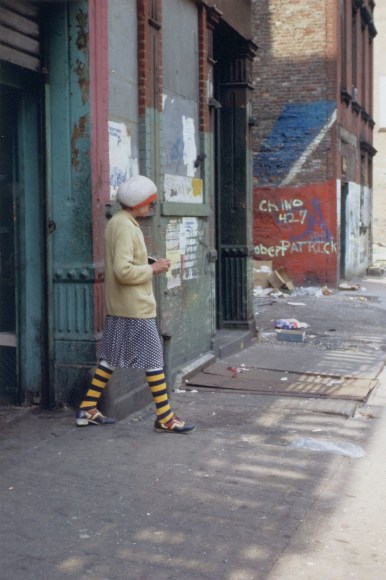 Color photo of an older woman in blue and yellow striped socks with a colorful mismatched outfit walking on a grimy street with graffiti in New York City.