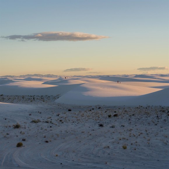 Color photo of sand dunes seen seen with the sun low in the sky, and four people walking in the distance.