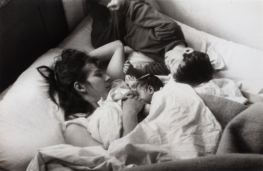 Black and white phto showing a mother nursing her infant child in bed, while an older child lies next to them.