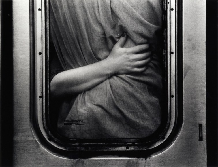 Black and white photo: Image of an arm clutching something draped in fabric, seen though a NYC subway door window.