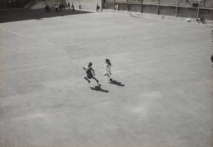 Black and white photo of two young girls running across a large paved lot, seen from a distance.
