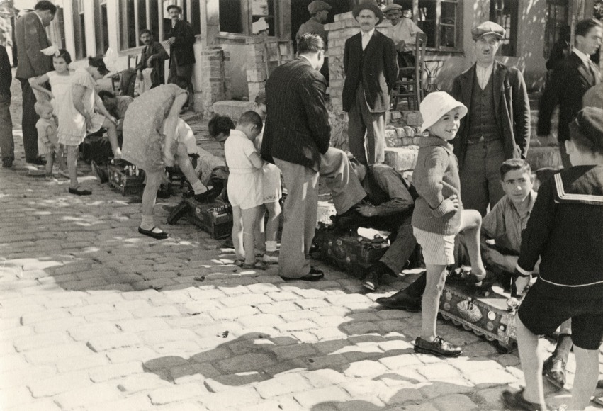 Black and white photo showing a young boy on a cobblestone street, among a group of people getting shoeshines.