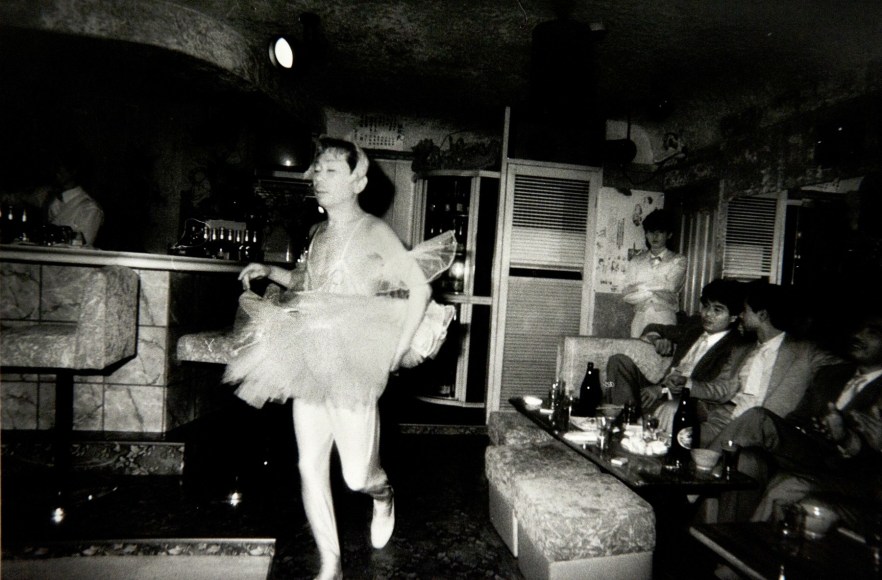 Black and white photo of a cross dressing man, wearing a ballerina outfit, parading in front of Japanese bar patrons who are seated and watching.