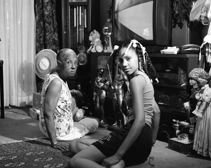 Black and thaw photo of young black woman taking a photo of herself and her grandmother, sitting on the floor of their family living room.