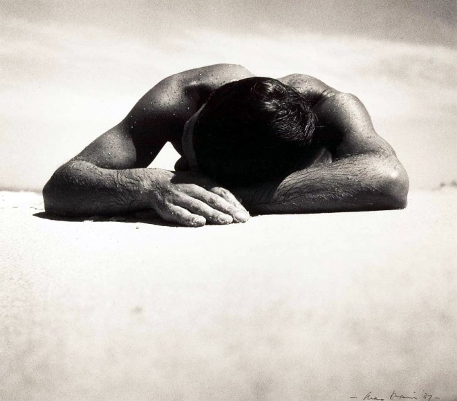 Black and white photo of a wet shirtless man sunbathing on a beach.