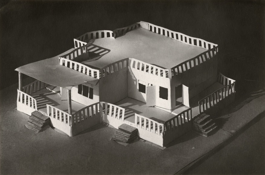 Black and white photo of small architectural model made out of paper&ndash;depicting a single story building with a fenced wrap around porch in front.