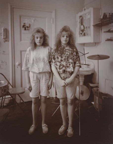 Posed black and white photo of twins sisters in a room with a drum kit.