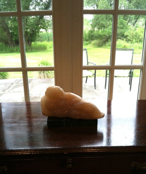 Small, translucent, pink tinted, white sculpture of a round female form lying propped up on its side.