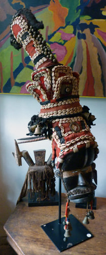 Wooden mask replicating a human profile. Resting on top of the mask is a red, black, and ivory/beige headdress, made of cloth, cowrie shells, fiber, bronze bells, glass beads, and kaolin. which extends far above the mask with colorful tassels that fall alongside the mask.