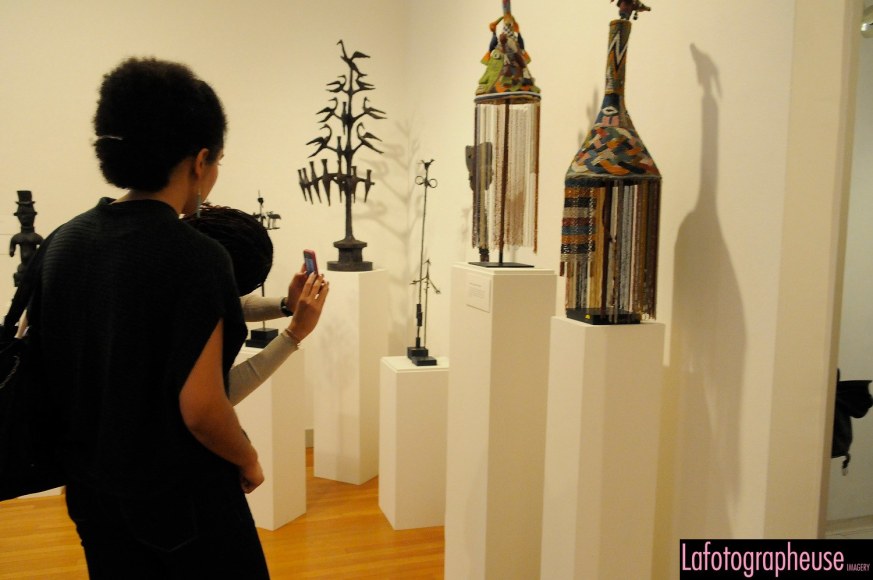 Two people stand on the left side of us looking at a colorful mask. The person furthest away from us has only their arms and head showing and is taking a picture. Both people have dark hair and the person standing closest to us is wearing a black, business casual outfit, and the other is wearing a grey long sleeve shirt.